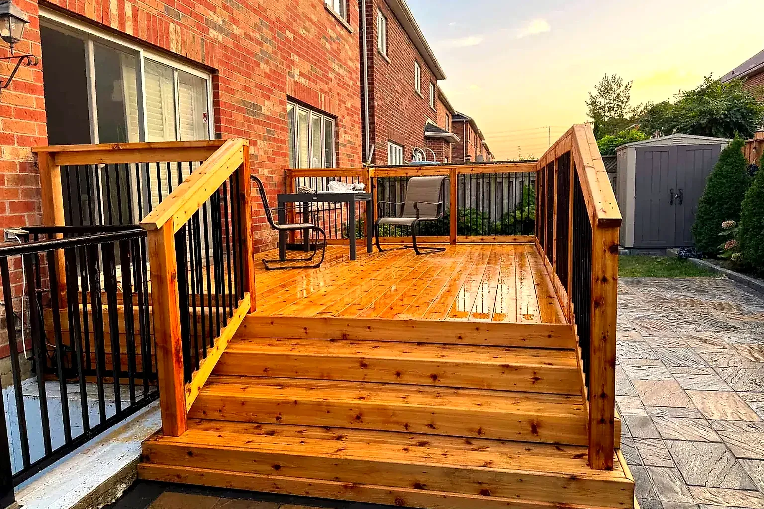 A new cedar deck with black metal balusters and wide stairs at the back of a brick home, at sunset